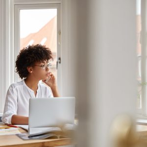 Photo of female entrepreneur speaks via cell phone, looks thoughtfully into window, analyzes paperwork from accounting department, checks database, sit at workplace, disposable coffee, laptop on desk